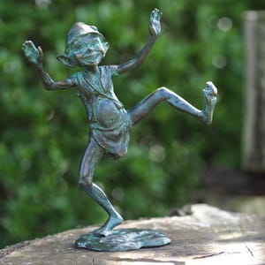 Könnte beinhalten: Bronzestatue eines tanzenden Elfen mit ausgestreckten Armen und einem erhobenen Bein. Der Elf trägt einen Hut und einen Umhang, die Statue hat eine grüne Patina. Die Skulptur steht auf einer natürlichen Holzoberfläche vor einem verschwommenen grünen Hintergrund.