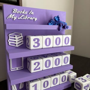 May include: A purple tiered shelf with the text "Books In My Library" and a small blue dragon figurine. The shelf features white cubes with purple numbers arranged in rows, alongside white icons representing books, a tablet, and headphones.