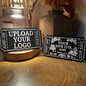 May include: Two black rectangular signs shaped like admission tickets with white raised lettering. The left sign reads UPLOAD YOUR LOGO, ADMIT ONE, and YOUR NAME. The right sign features floral graphics, a cup, books, and reads YOUR BOOK CLUB NAME, BOOK CLUB, ADMIT ONE, and YOUR NAME.