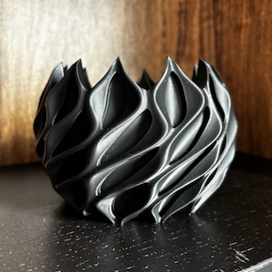 May include: A black, 3D-printed decorative bowl with a unique, wave-like design. The bowl has a series of curved, vertical ridges that create a textured surface. The bowl is likely made of plastic and is suitable for home decor.