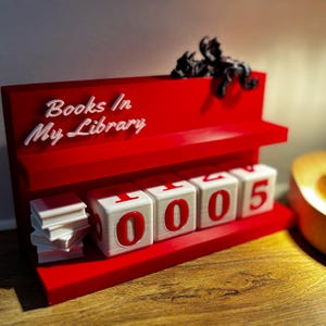 May include: A red and white 3D-printed book counter with the text "Books In My Library." The counter displays "1005" with white cubes and red numbers. A small stack of books and a black figurine are also present.