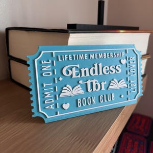 Könnte beinhalten: Ein blau-weißes 3D-gedrucktes Schild in Ticketform für den "Endless TBR Book Club". Auf dem Schild steht "Lifetime Membership" und "List Lover". Das Design umfasst Symbole für offene Bücher und die Worte "Admit One".