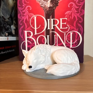 May include: A white wolf figurine in a sleeping pose, resting on a gray, circular base. The figurine is in front of a book with the title "Dire Bound" in large white letters against a red background. The figurine is a decorative item.