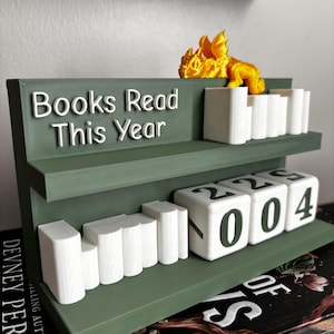 May include: A green bookshelf with the text "Books Read This Year" in white. The shelf has a dragon figurine and white book-shaped markers. The bottom shelf displays the number "2004".