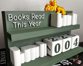 Puede incluir: Una estantería verde con el texto "Books Read This Year" en blanco. La estantería tiene una figura de dragón dorado y marcadores blancos con forma de libro. La parte inferior muestra el número "2004".