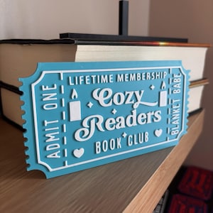 May include: A light blue, ticket-shaped sign with white lettering that reads "Lifetime Membership Cozy Readers Book Club." The sign has a decorative edge and is placed in front of a stack of books.