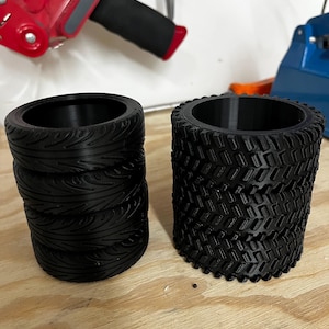May include: Two black 3D-printed tire-shaped containers. One has a smooth, curved tread pattern, while the other features a more aggressive, blocky tread design. Both are cylindrical and appear to be empty inside. They are sitting on a wooden surface.