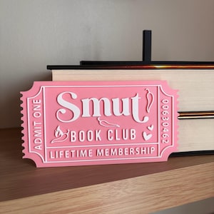 May include: A pink ticket-shaped sign with the words "Smut Book Club Lifetime Membership" in white. The sign has a decorative border and includes small chili pepper and heart graphics. The ticket reads "Admit One" and has a serial number.