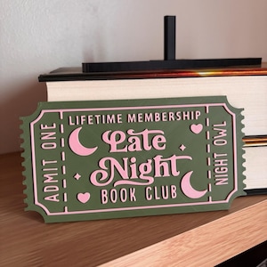 May include: A green and pink sign in the shape of a ticket that reads "LIFETIME MEMBERSHIP Late Night BOOK CLUB". The sign has a scalloped edge and features crescent moons, hearts, and stars.