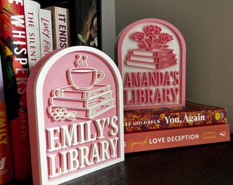 Puede incluir: Dos letreros decorativos rosas y blancos para bibliotecas. Uno dice "Emily's Library" y presenta una taza de café, libros y una flor. El otro dice "Amanda's Library" y presenta rosas y libros. Se ven libros en el fondo.