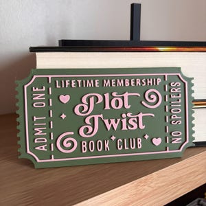 Peut inclure: Panneau vert et rose en forme de billet imprimé en 3D pour le "Plot Twist Book Club". Le panneau indique "Adhésion à vie" et "Admettez-en un", avec les mots "Plot Twist Book Club" dans une police décorative. Le panneau dit aussi "Pas de spoilers."