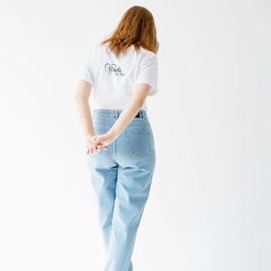Peut inclure: Un t-shirt blanc avec l'inscription "Bride to Be" en noir, associé à un jean bleu clair. La personne est vue de dos, les mains jointes. La tenue est complétée par des baskets blanches. Le fond est blanc uni.