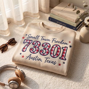 May include: A beige sweatshirt with the text "Small Town Freedom 73301 Austin, Texas" in navy blue and red plaid. The sweatshirt is folded on a white surface, with sunglasses and headphones nearby.