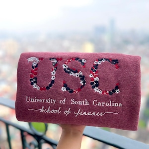 Może przedstawiać: Bordowy prostokątny przedmiot z haftowanymi literami "USC" we wzór kwiatowy. Poniżej liter znajduje się napis "University of South Carolina School of Finance" białą nicią.