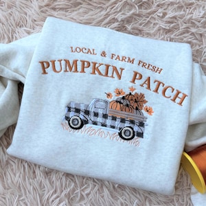 May include: A light gray sweatshirt with an embroidered design. The design features a black and white plaid truck carrying pumpkins, with the text "LOCAL & FARM FRESH PUMPKIN PATCH" in orange thread. The sweatshirt is folded on a soft, textured surface.