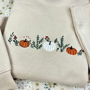 Peut inclure: Sweat-shirt beige avec broderie sur le thème de l'automne. Il présente trois citrouilles orange et blanches, ainsi que du feuillage vert et des baies rouges. Le motif est centré sur la poitrine.