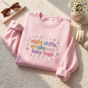 Könnte beinhalten: Hellrosa Sweatshirt mit dem gestickten Text "night shifts, scrubs and baby hugs" in bunten Buchstaben. Das Design beinhaltet gestickte Gänseblümchen und Sterne. Das Sweatshirt hat gerollte Ärmel und einen Rundhalsausschnitt.