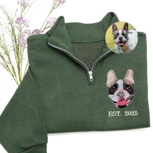 May include: A dark green sweatshirt with a quarter-zip closure and a detailed embroidered French Bulldog design. The design includes the text "EST. 2025". A small image of a French Bulldog is also present.
