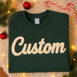May include: A dark green sweatshirt with the word "Custom" in a cream-colored, textured font. The sweatshirt is folded on a white, fluffy surface, with Christmas ornaments and candy canes in the background.