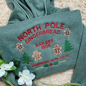 Może przedstawiać: Zielona bluza z kapturem z haftowanym napisem "NORTH POLE GINGERBREAD BAKERY 1909". Projekt zawiera piernikowych ludzików i choinki. Fraza "HANDMADE WITH LOVE" jest również wyhaftowana.