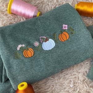 Peut inclure: Un sweat-shirt vert avec des broderies de citrouilles et des motifs floraux. Le motif présente trois citrouilles orange et bleu clair, avec des fleurs roses et blanches et des feuilles vertes. Le fond comprend des bobines de fil rose et orange.