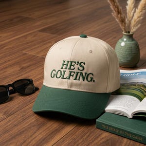 May include: A beige and green baseball cap with the text "HE'S GOLFING." The cap has a green brim and button. Sunglasses and a golf magazine are next to the cap.