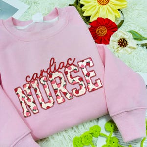 May include: A pale pink sweatshirt featuring the embroidered words "cardiac NURSE". The word "cardiac" is in red script, while "NURSE" is filled with a strawberry pattern. It has a crew neck and long sleeves.