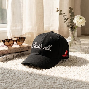 May include: Black baseball cap with the embroidered phrase "That's all." in white script. A red high-heeled shoe graphic is on the side. Tortoiseshell sunglasses and books are in the background.