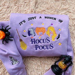 Pode incluir: Um moletom lilás com a frase bordada "IT'S JUST A BUNCH OF HOCUS POCUS" e um gráfico de três bruxas. O design inclui uma vassoura, morcegos e estrelas. Uma aranha preta e uma lanterna de abóbora também são visíveis.