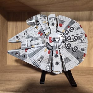 May include: A detailed model of the Millennium Falcon spaceship constructed from grey, white, and dark grey plastic building bricks. The circular vessel features a cockpit, a central gun turret, and small red accents, mounted on a black stand.