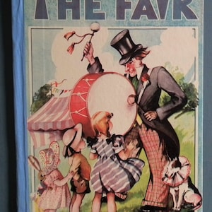 May include: A vintage children's book cover with the title "THE FAIR" in blue letters. The cover features a man in a top hat playing a drum, three children, and a dog. The book is by Mary Brough.