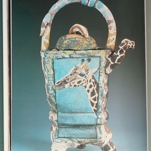 Könnte beinhalten: Eine Teekanne aus Keramik mit einem Giraffen-Design. Die Teekanne hat einen blauen und weißen Körper mit einem Giraffenkopf, der aus der Vorderseite herausragt. Die Teekanne hat einen Griff in Form eines Giraffenhalses und einen Ausguss in Form eines Giraffenkopfes.