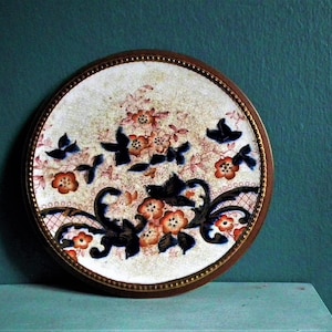 Peut inclure: Assiette décorative ronde avec un bord marron et un motif floral. L'assiette présente un fond blanc avec des motifs floraux orange et noirs et des silhouettes d'oiseaux noirs. L'assiette est probablement en céramique ou en porcelaine.