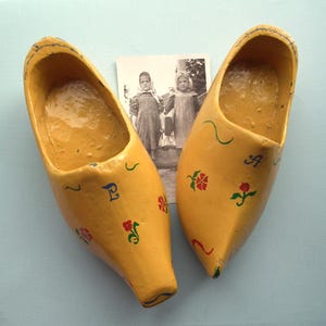 May include: A pair of yellow wooden clogs with painted floral and letter designs. A vintage black and white photograph of two girls is behind the clogs. The clogs are a traditional style, likely for casual wear.