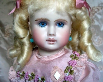 May include: A porcelain doll with blonde curly hair tied with pink ribbons, bright blue eyes, and dangling turquoise earrings. The doll wears a pink dress with lace trim, green and pink floral embroidery, and a central white diamond-shaped accent.