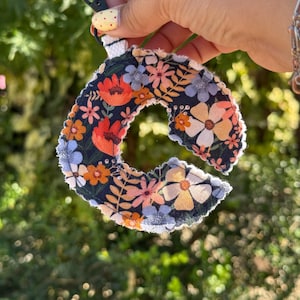 Könnte beinhalten: Ein Schlüsselanhänger mit dem Buchstaben "C" aus Stoff in Marineblau mit einem schwarzen Clip. Der Buchstabe hat ein Blumenmuster in Orange, Pfirsich und Weiß. Die Kanten sind ausgefranst und wirken handgemacht.