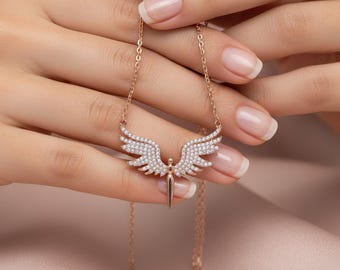 May include: A rose gold necklace with a pendant shaped like wings, adorned with small, sparkling crystals. The necklace is held by a person with manicured nails, set against a soft pink backdrop.