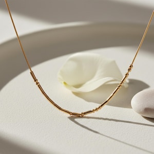 Könnte beinhalten: Eine zarte goldene Halskette mit linearem Design, die kleine zylindrische und kugelförmige Perlen aufweist. Die Halskette wird auf einer weißen Oberfläche mit einem weißen Blütenblatt und einem kleinen Stein präsentiert, was eine minimalistische Ästhetik erzeugt.