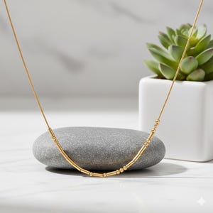 May include: A delicate gold necklace with a linear design. The necklace features a thin chain and a series of small, cylindrical and spherical beads. The necklace is displayed on a gray stone, with a succulent plant in the background.