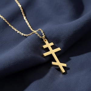 Könnte beinhalten: Eine goldfarbene Halskette mit einem Anhänger in Form eines russisch-orthodoxen Kreuzes. Der Anhänger hat drei horizontale Balken und einen schrägen unteren Balken. Die Halskette ist zart und goldfarben, vor einem dunkelblauen Stoffhintergrund.