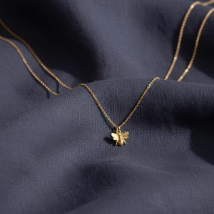 Może przedstawiać: Delikatny, złoty naszyjnik z małą zawieszką w kształcie pszczoły. Zawieszka pszczoły ma szczegółowe skrzydła i korpus. Łańcuszek naszyjnika jest cienki i wisi na tle ciemnoniebieskiej tkaniny.