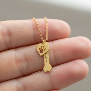 Op de afbeelding: Een goudkleurige ketting met een hartvormige hanger met een uitgesneden pootafdruk en een botvormige bedel. Het woord "Buddy" is gegraveerd op de botbedel. De ketting wordt getoond tegen een wazige achtergrond.