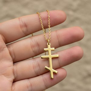 May include: A gold-colored Orthodox cross pendant necklace. The pendant features a three-barred cross design, suspended from a delicate chain. The necklace is held against a blurred background.