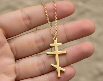 May include: A gold-colored Orthodox cross pendant necklace. The pendant features a three-barred cross design, suspended from a delicate chain. The necklace is held against a blurred background.