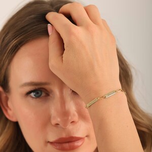 May include: A gold-colored bracelet with two rectangular nameplates, each engraved with a name. The bracelet is linked with small, rectangular links. The names "Aurora" and "Maya" are visible on the nameplates.