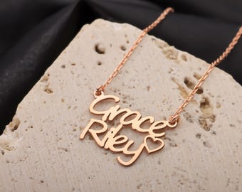 May include: Rose gold necklace featuring the names "Grace" and "Riley" in a cursive font. A small heart design is placed at the end of the name "Riley". The necklace is displayed on a textured, light-colored surface.