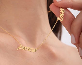 May include: A gold-colored necklace with the word "peace" in a cursive font. A separate charm with the word "love" is held up by a hand. The necklace is worn around a person's neck.