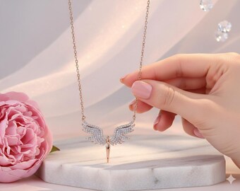 May include: A rose gold necklace featuring a crystal-encrusted angel wing pendant. The necklace is held by a hand with pink nail polish. A pink peony flower is in the background. The necklace is displayed on a white surface.