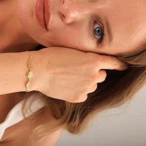 Op de afbeelding: Een goudkleurige armband met delicate vleugel- en bloembedels. De armband is gemaakt van een fijne ketting en wordt om de pols gedragen. De bedels zijn ingewikkeld gedetailleerd en geven de sieraden een vleugje elegantie.