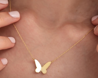 May include: A delicate gold-colored necklace featuring a butterfly pendant. The butterfly has a simple, elegant design with two wings. The necklace is displayed against a skin-toned background.